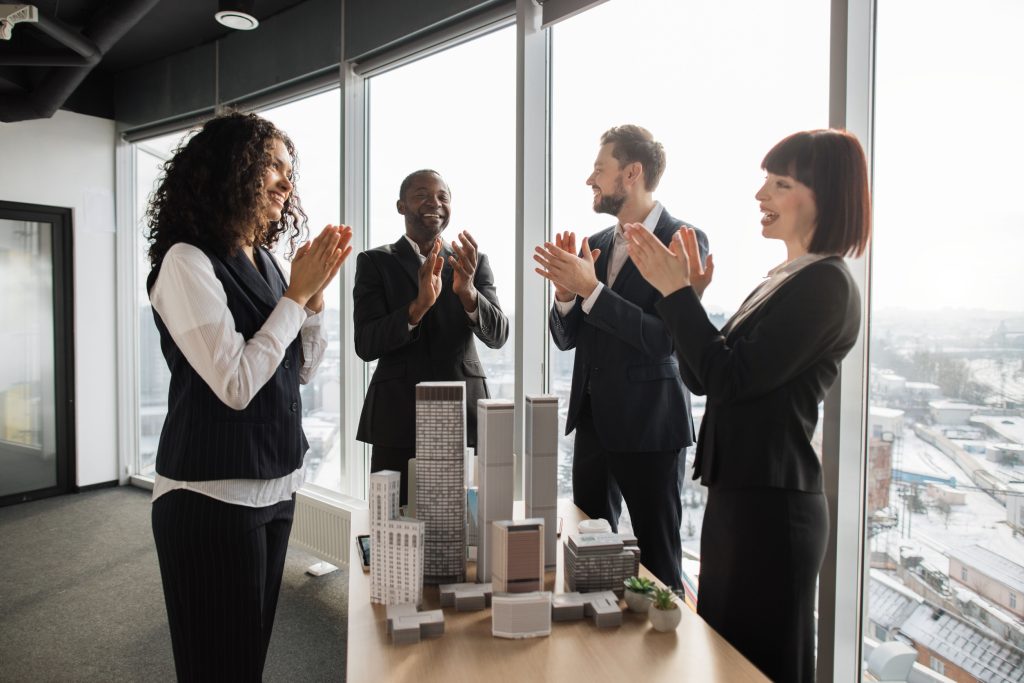 Good results, congratulation and appreciation concept. Satisfied diverse business men and women in formal wear, standing near table with skyscraper building maquette in conference hall and applauding
